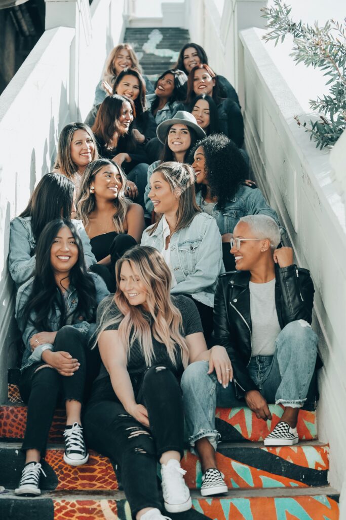 Group of diverse women sitting on steps together, smiling and laughing, symbolizing different stages of womanhood and shared experiences.